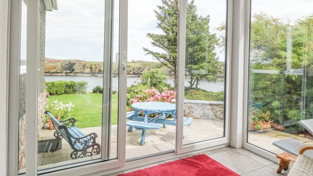A room with a sliding glass door leading to an outdoor patio with a table and chairs, a red rug, and a view of a lake and greenery. - The Headland (Amlwch)