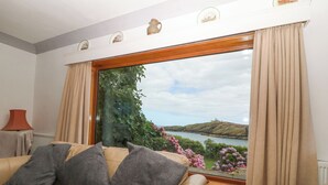 A living room with a beige sofa, grey cushions, a wooden-framed window, and a view of a lake and hills. - The Headland (Amlwch)