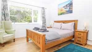 A bedroom with a wooden bed, a chair, a nightstand, a blue rug, and a window with curtains. - The Headland (Amlwch)