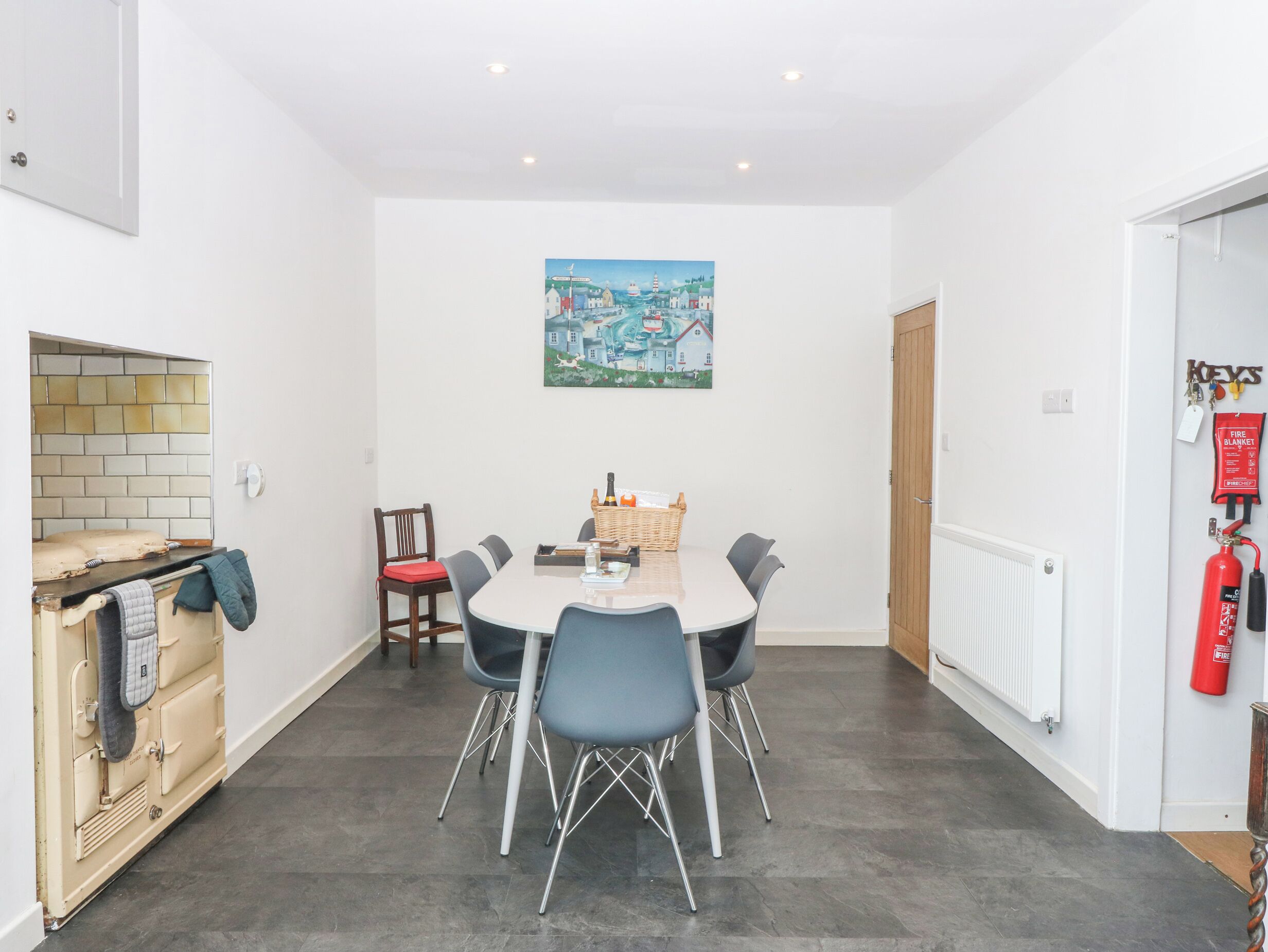 A dining area with a white table, six chairs, a painting on the wall, a fire extinguisher, and a vintage oven.