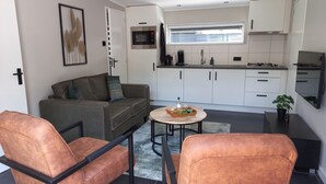 Ein kompakter Wohnbereich mit Sofa, zwei Sesseln, einem kleinen runden Tisch, einer Küchenzeile mit weißen Schränken, einer Mikrowelle und einem Fenster.