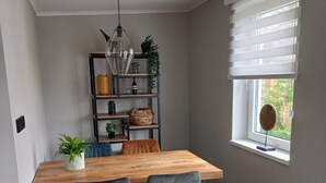 Ein Essbereich mit einem Holztisch, zwei Stühlen, einer Pendelleuchte, einem Regal mit Dekoration und einem Fenster mit Jalousien.