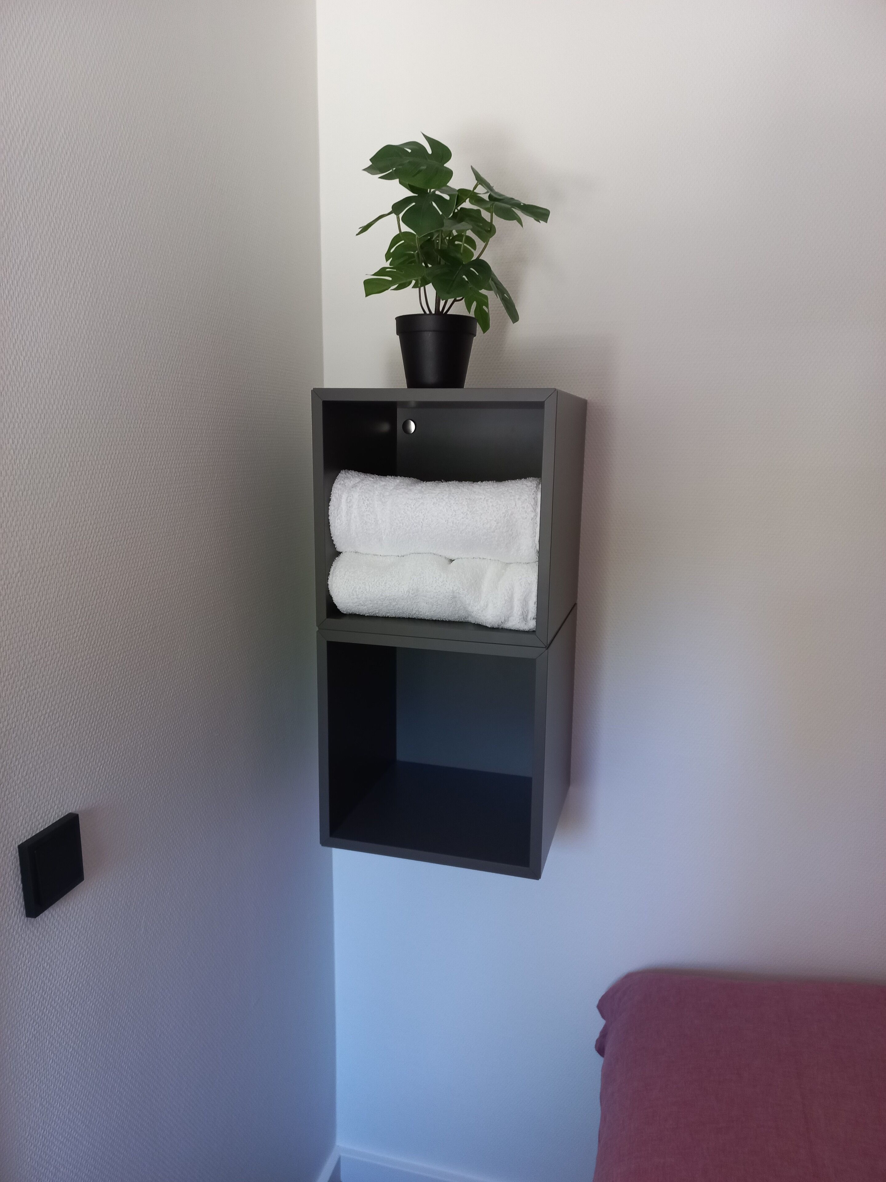 A corner shelf unit with two shelves, one holding a potted plant and the other holding folded towels.