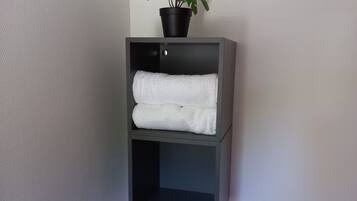 A corner shelf unit with two shelves, one holding a potted plant and the other holding folded towels.