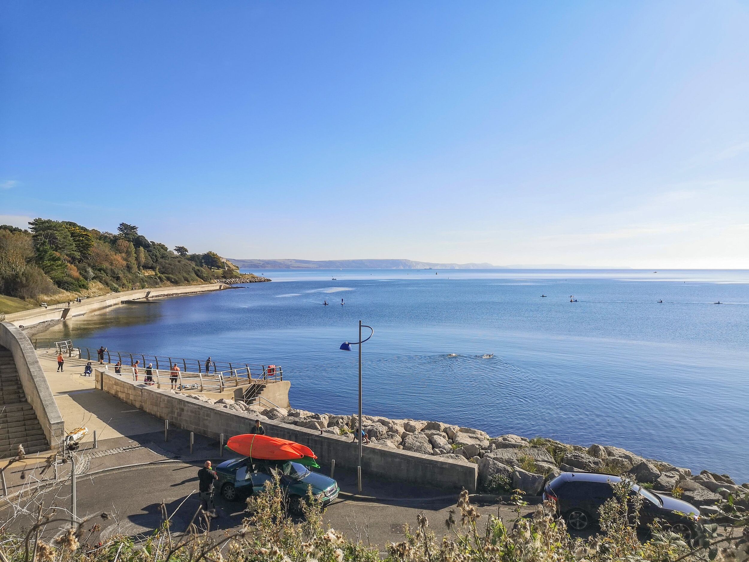 A coastal area with a paved walkway along the water, a parking area, and a view of the sea.