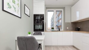 A modern kitchen with white cabinets, a grey chair, a small potted plant, and a window with a view of buildings.