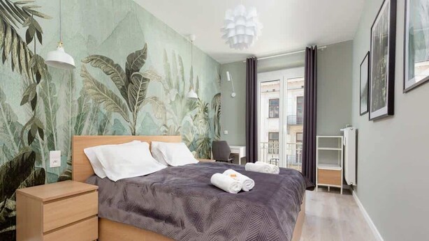 A bedroom with a bed, a nightstand, a wall mural, a window with curtains, and a radiator.
