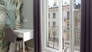 A modern apartment with a white desk, a grey chair, a large window with a view of buildings and a crane, and a tropical mural on the wall.