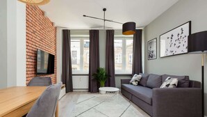 A modern living room with a brick wall, a flat-screen TV, a grey sofa, a dining table with chairs, and a large window with curtains.