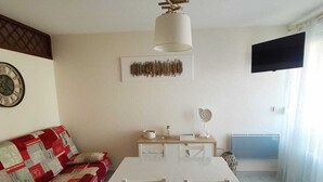 Zona pranzo con tavolo e sedie bianche, una ventola a soffitto, TV a parete e un divano con motivo decorativo.