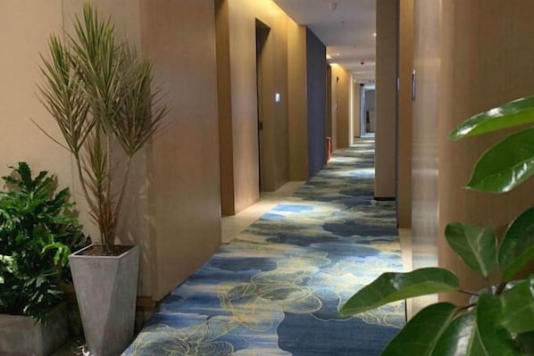 Un couloir d’hôtel avec un tapis à motifs, des plantes en pot et un long couloir bordé de portes de chaque côté.