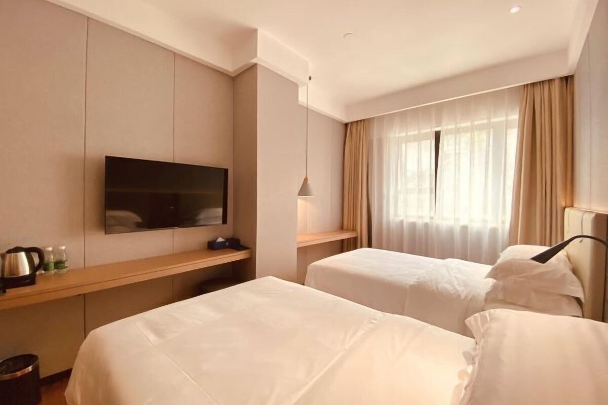 A hotel room with two beds, a TV mounted on the wall, a bedside table with a kettle, and a window with curtains.