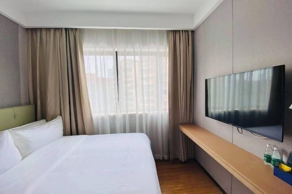 A hotel room with a bed, a TV, a window with curtains, and a wooden bench.