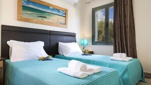 A hotel room with two beds, each with a teal bedspread and white pillows. There is a painting of a beach scene on the wall, a window with curtains, and a bedside lamp. - Rose Villa in Svoronata (Kefalonia)