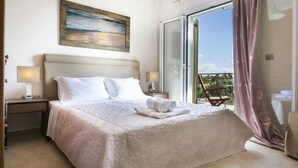 A hotel room with a bed, a painting on the wall, a balcony with a chair, and a view of the outdoors. - Rose Villa in Svoronata (Kefalonia)