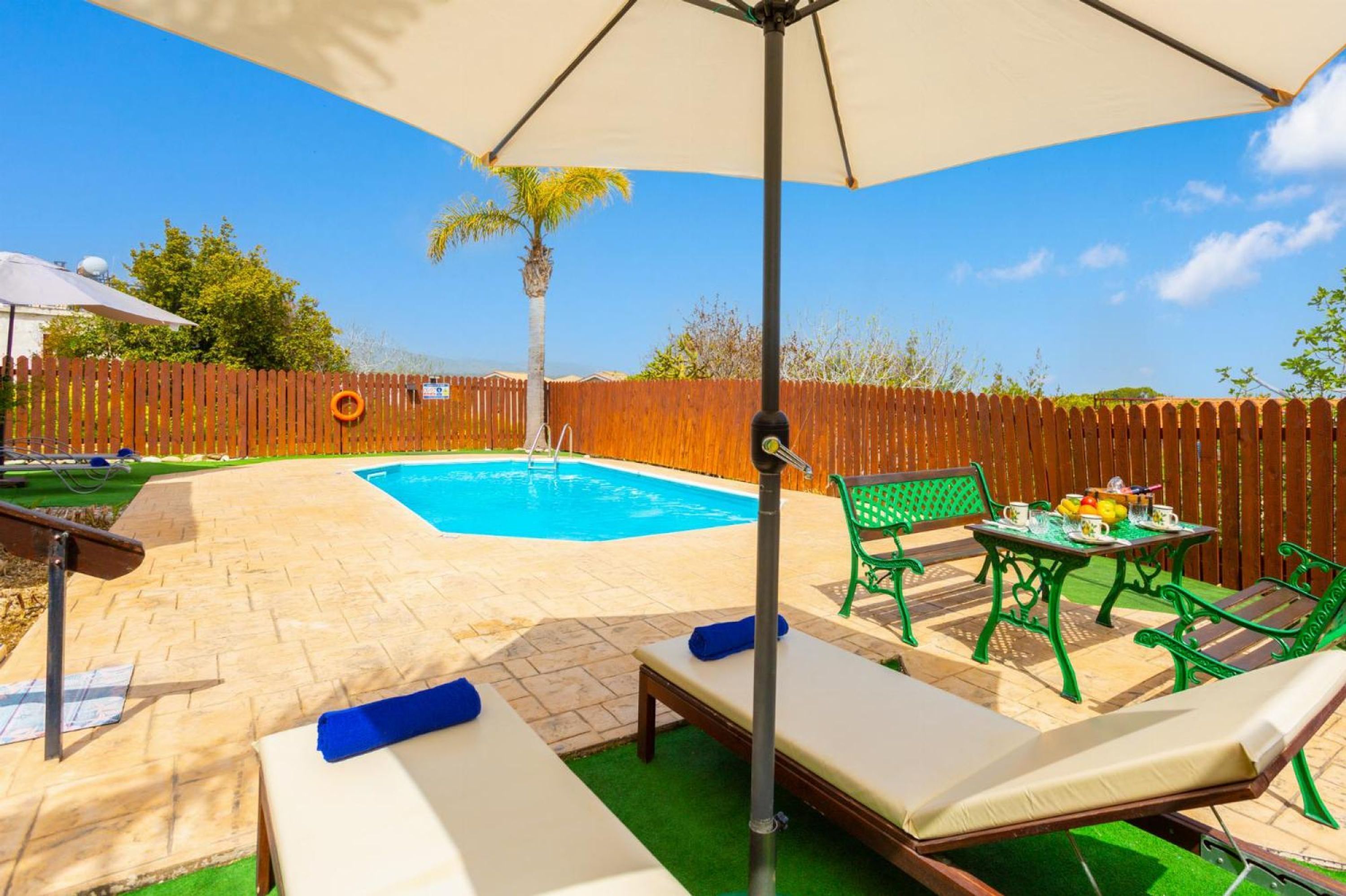 Un jardin avec une piscine, des chaises longues, une table avec des boissons et une clôture en bois.