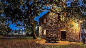 A stone building with a large tree in front, a picnic table, and a well-lit entrance.