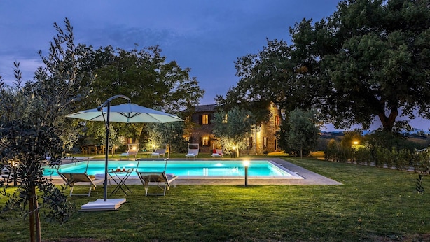 A swimming pool with lounge chairs and an umbrella, surrounded by a well-maintained lawn and trees, with a stone building in the background.