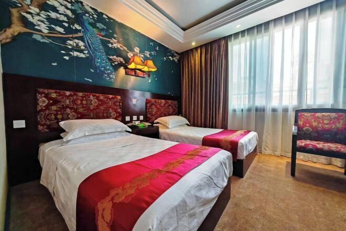 A hotel room with two beds, a decorative wall mural, a large window with curtains, and a chair.