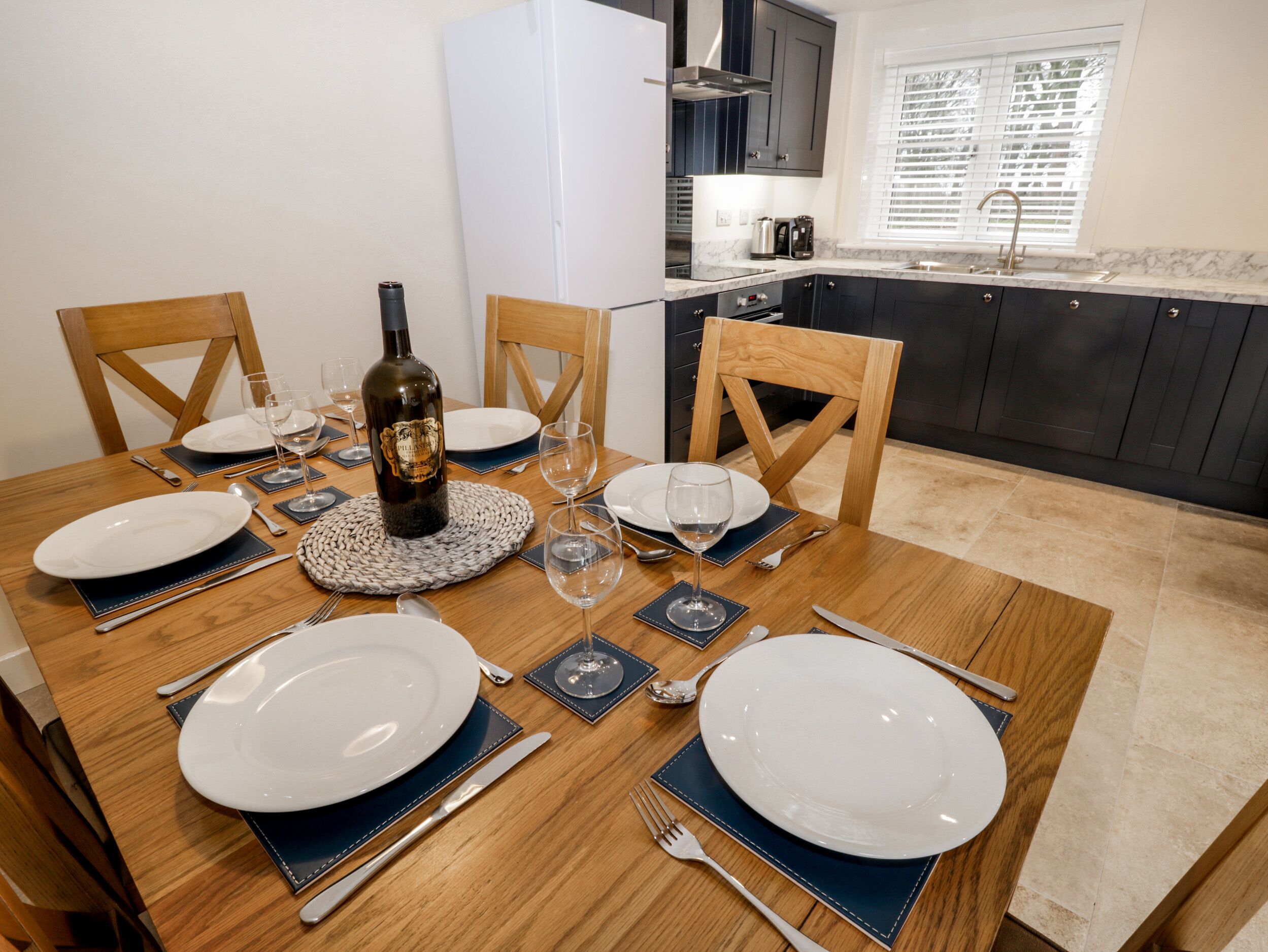 Una mesa de comedor dispuesta con platos, cubiertos y una botella de vino, rodeada de sillas en una cocina moderna con armarios oscuros y una ventana con persianas.