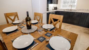 Una mesa de comedor dispuesta con platos, cubiertos y una botella de vino, rodeada de sillas en una cocina moderna con armarios oscuros y una ventana con persianas.