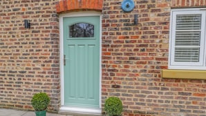 A brick building with a green door, two potted plants, and a window with blinds. - The Grooms Cottage (York)