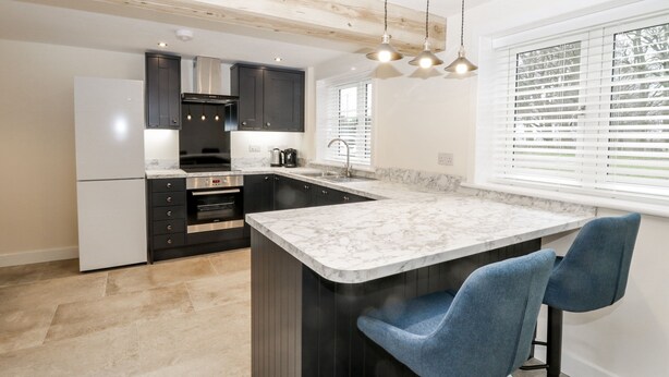 A modern kitchen with dark cabinets, a marble countertop, and a built-in oven. There are two blue bar stools and a window with blinds. - The Grooms Cottage (York)