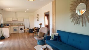 Una sala moderna con un sofá azul, una mesa de centro redonda y una silla de mimbre. Hay una zona de cocina con microondas, tetera y una mesa de comedor con sillas.