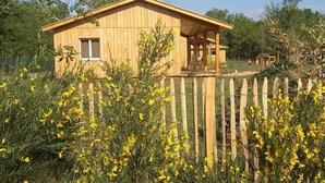Una cabaña de madera con un porche, rodeada por una valla de madera y plantas de flores amarillas.
