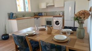Una cocina moderna con armarios blancos, microondas integrado, lavavajillas y lavadora. Hay una mesa de comedor con sillas, un florero con flores secas y una planta en maceta.