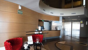 Un hall d’hôtel avec un mur en bois, un comptoir d’accueil, un panneau indiquant « SORTIE » et deux chaises rouges sur lesquelles repose un vase en verre, posé sur une petite table.
