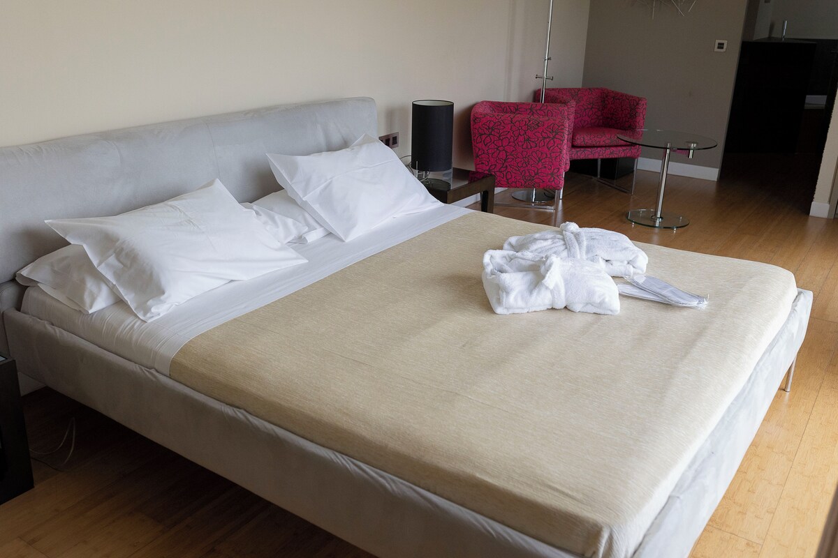 Una cama bien hecha con sábanas blancas y una manta color beige, una silla roja con estampado y una mesita redonda con una lámpara.