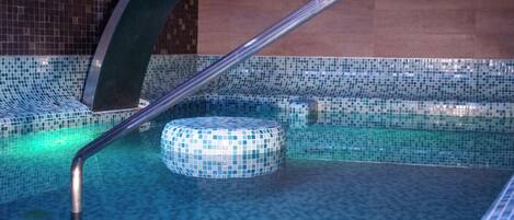A section of a pool with mosaic tiles, a metal handrail, and a circular seating area.