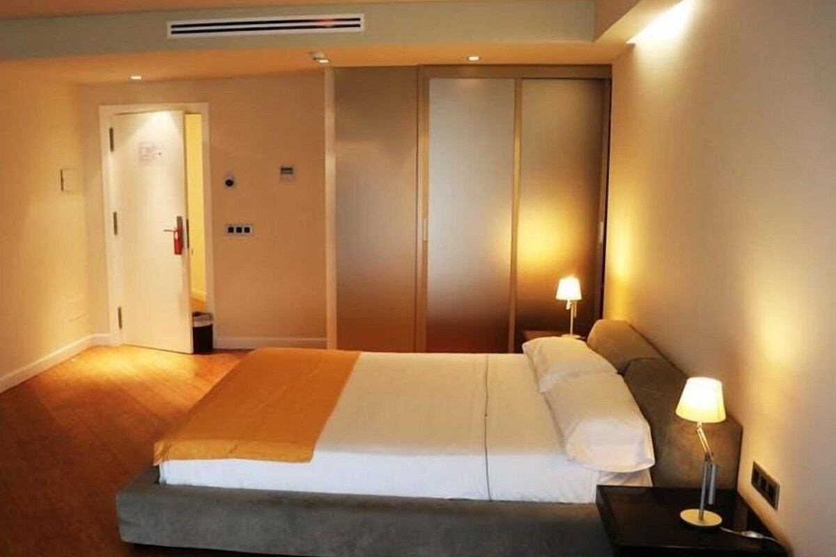 Una habitación de hotel con cama, lámparas de noche, un armario y una puerta.
