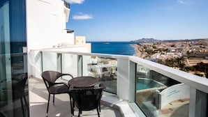 Un balcon doté de chaises en osier et d’une rambarde en verre, offrant une vue sur une ville côtière et la mer.