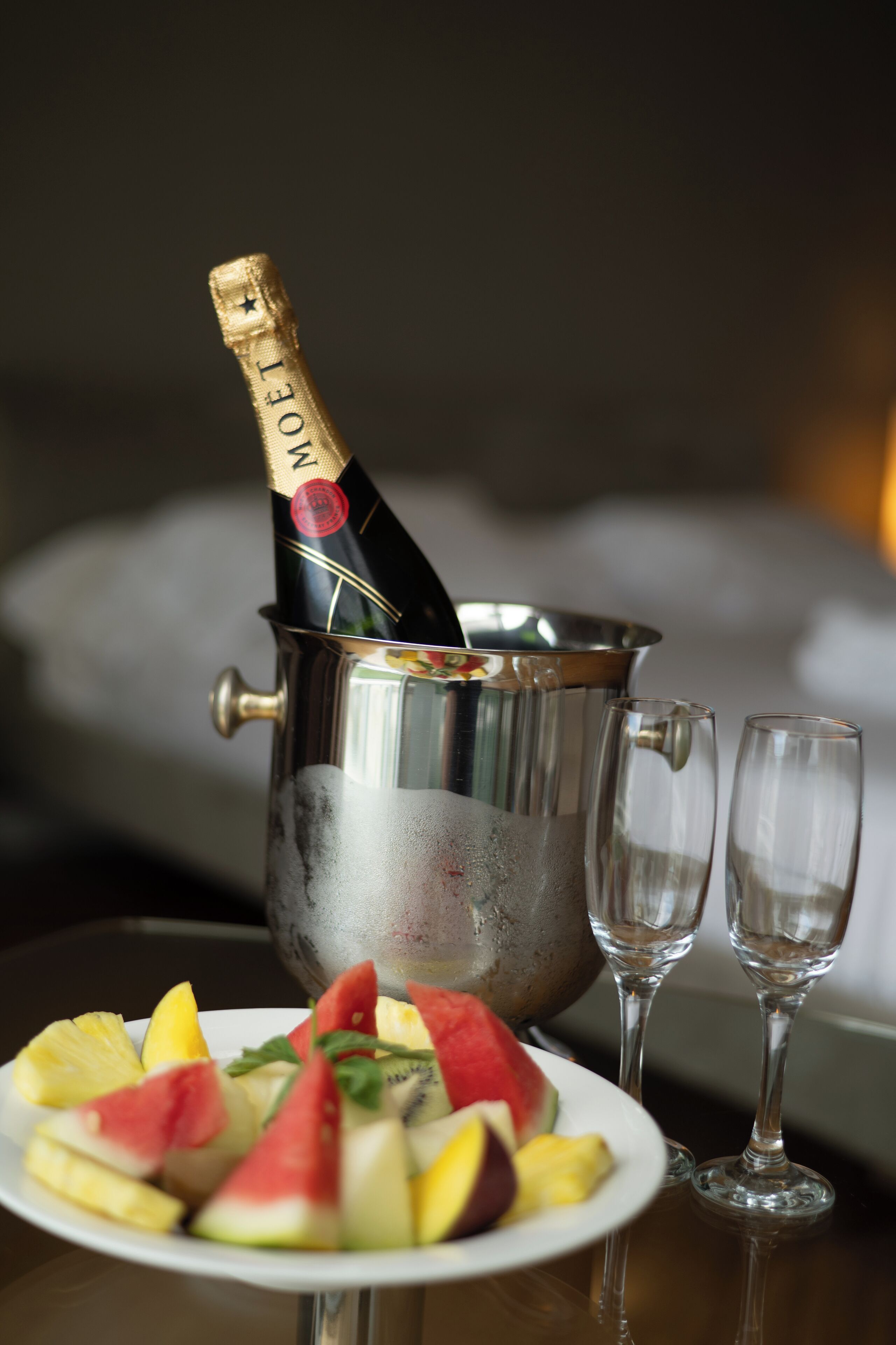 Eine Champagnerflasche in einem Metallkübel, zwei leere Gläser und eine Schale mit Fruchtschnitten.