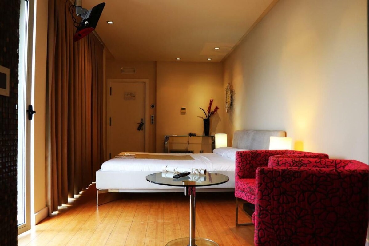 Una camera d'albergo con un letto, un tavolino rotondo con un telecomando, una sedia rossa e una lampada.