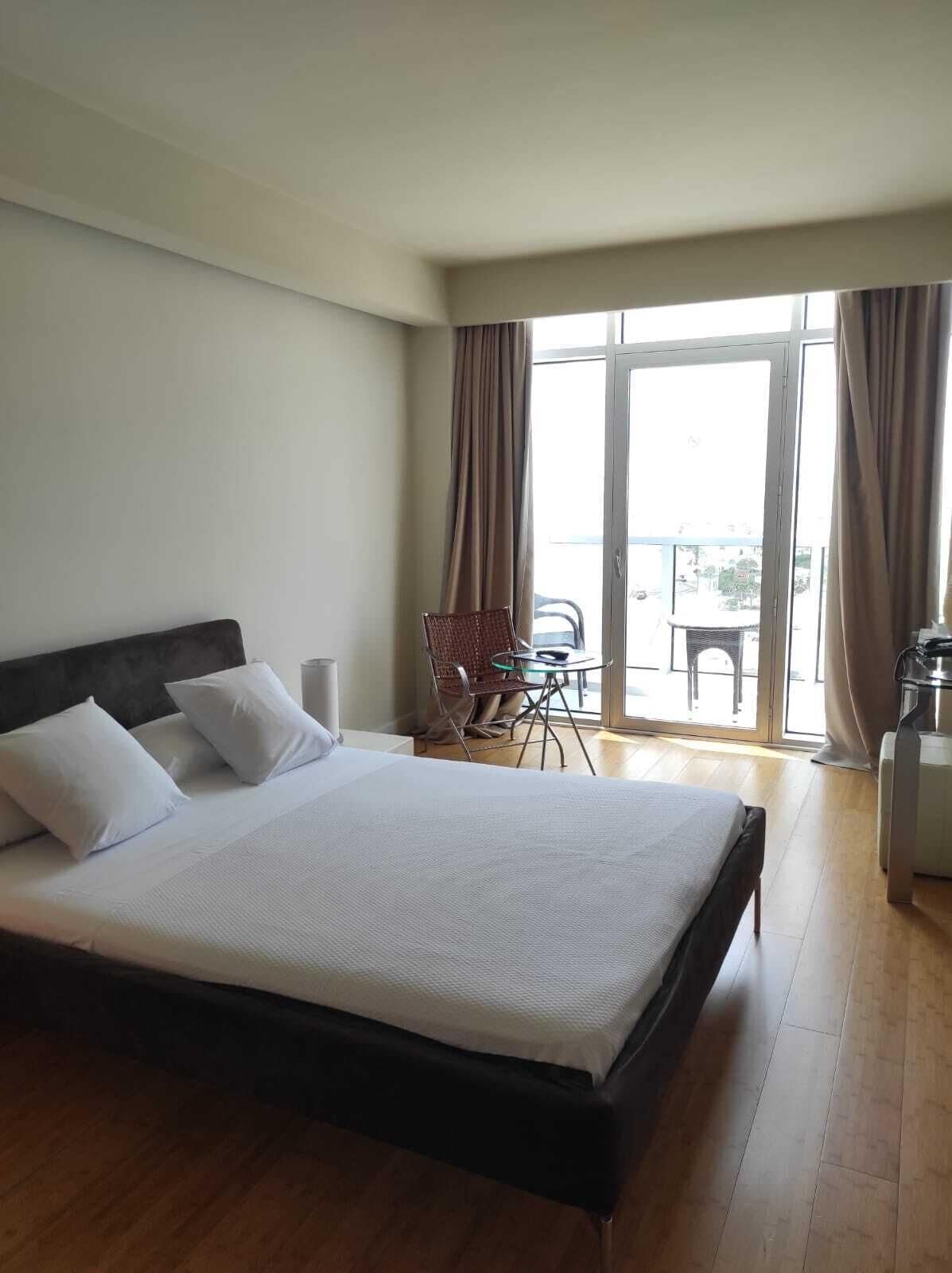 A hotel room with a large bed, a chair, a small table, and a balcony with curtains.