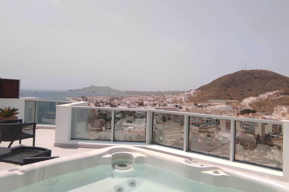 A rooftop terrace with a hot tub, a chair, and a view of a coastal town and hills.