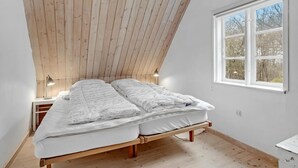 Quarto com teto inclinado, cama com roupa de cama branca, abajur e janela com vista para árvores.