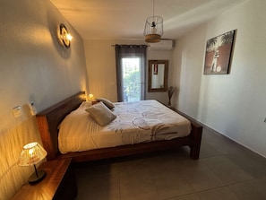 A bedroom with a bed, bedside lamps, a window with curtains, a hanging light fixture, and a wall-mounted mirror. - 2-room apartment with a veranda and a small garden (La Savane)