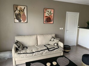 A living room with a sofa, two cushions, a coffee table with a decorative item, and two wall-mounted paintings. - 2-room apartment with a veranda and a small garden (La Savane)