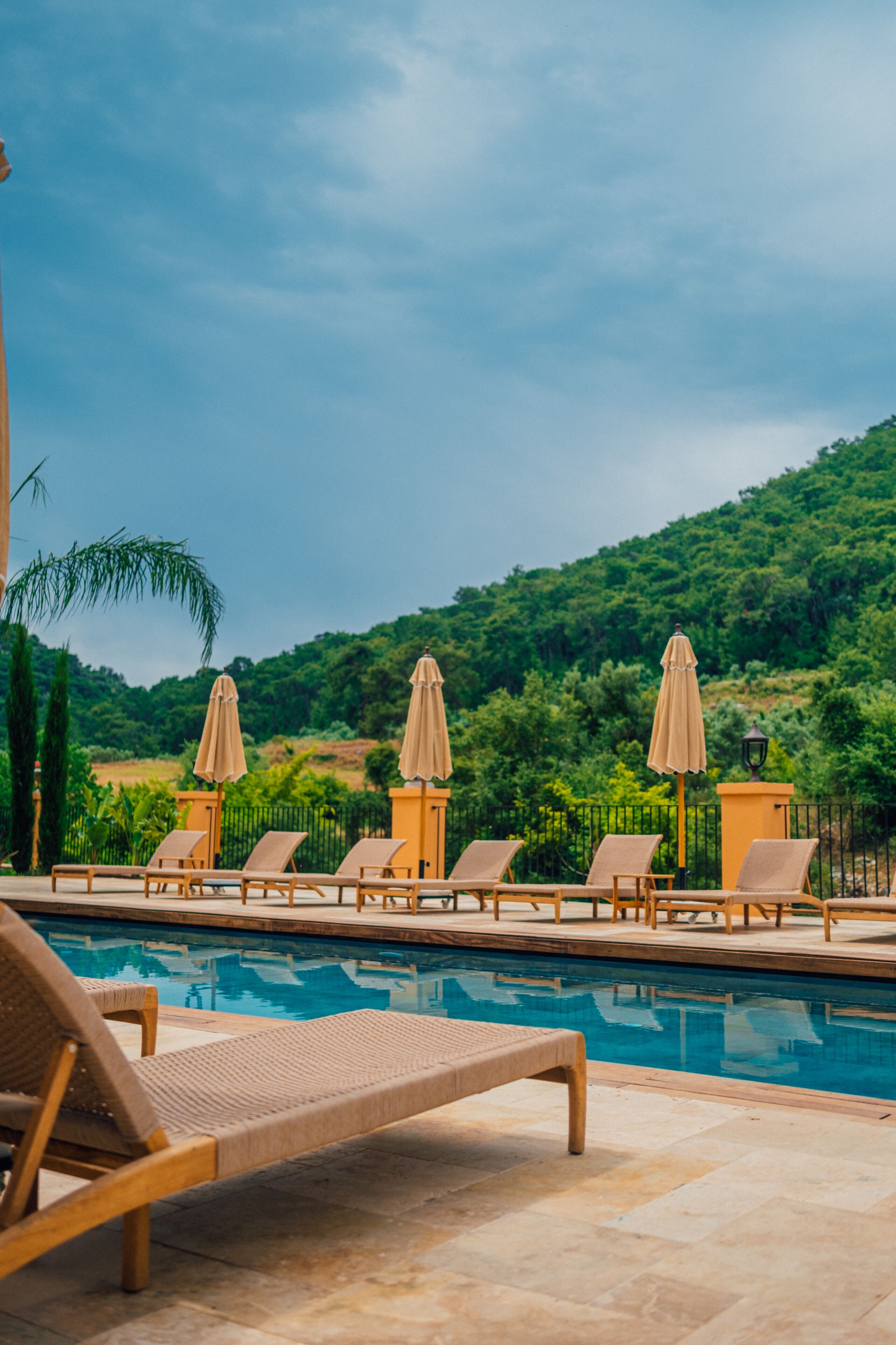 Zona junto a la piscina con sillones de descanso y sombrillas, rodeada de vegetación y un paisaje montañoso.