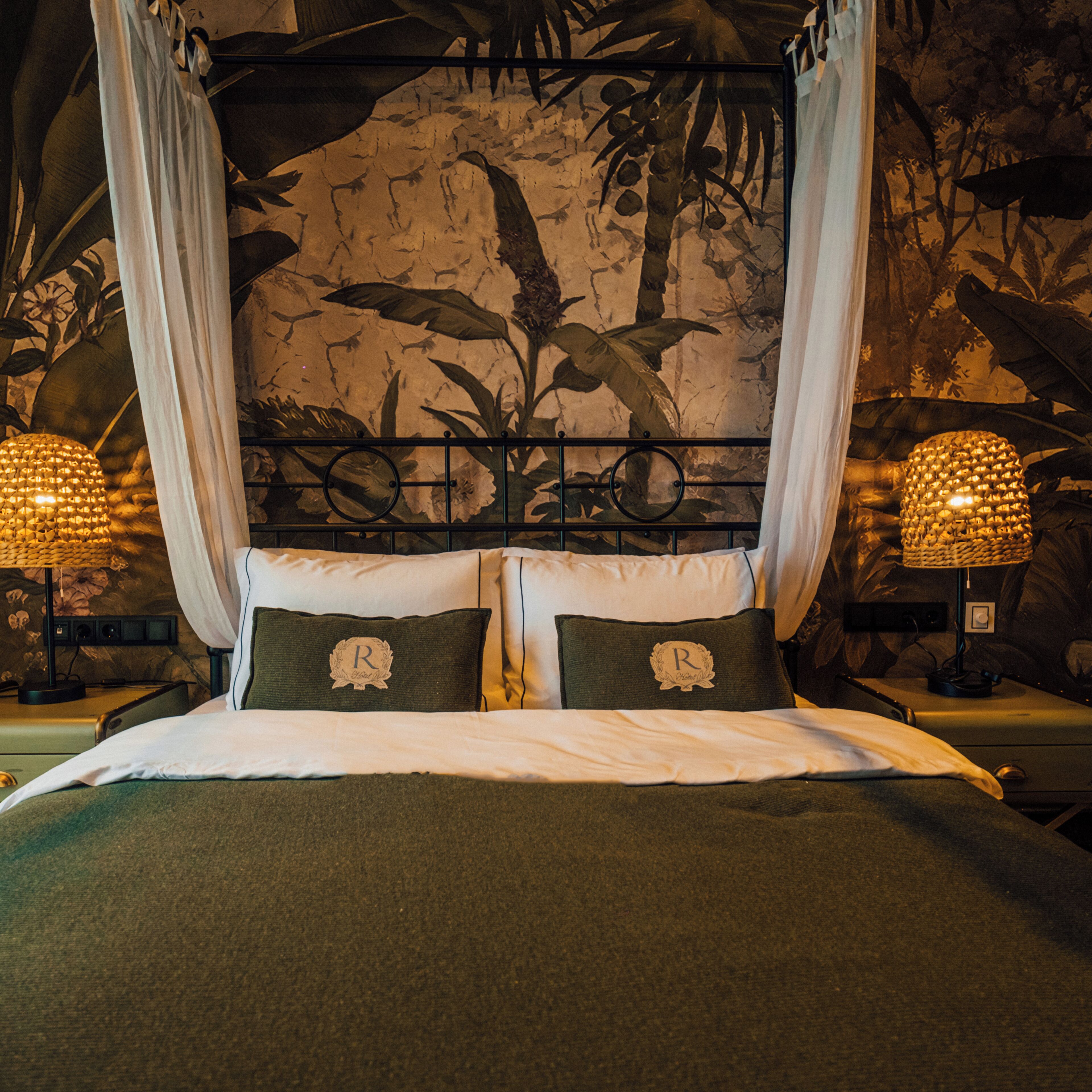 Una cama con dosel, dos almohadas con fundas con monogramas y una pared con un mural temático de naturaleza.