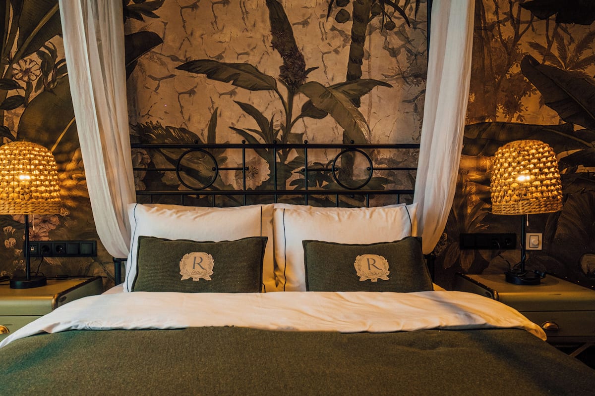 A bed with a canopy, two pillows with monogrammed covers, and a wall mural of foliage.