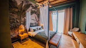 A bedroom with a canopy bed, a nightstand, a wooden bench, a desk, and a mural of tropical foliage.
