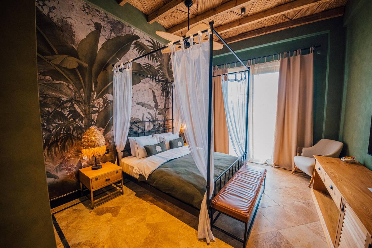 A bedroom with a canopy bed, a nightstand, a wooden bench, a desk, and a mural of tropical foliage.