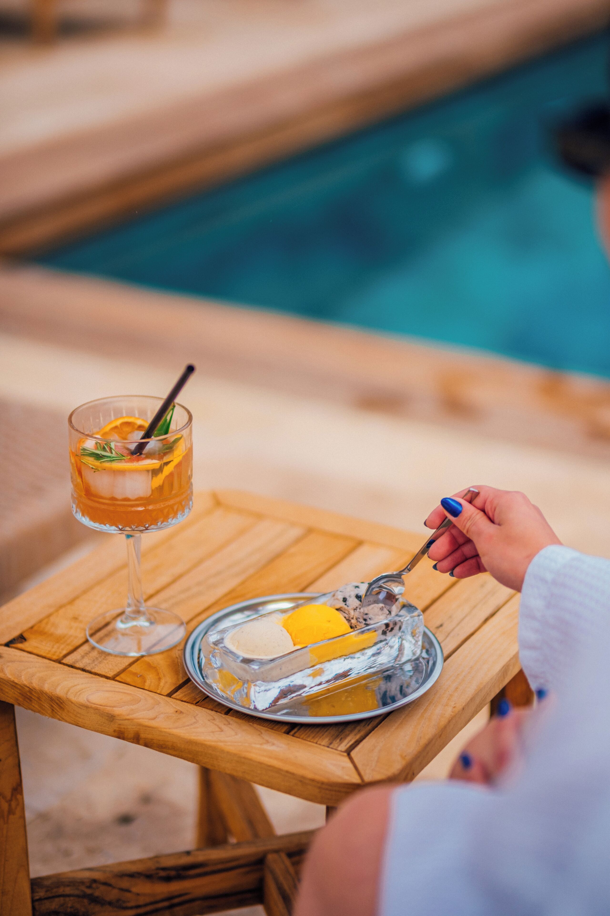 A wooden table with a glass of a beverage, a spoon, and a plate with a dessert.