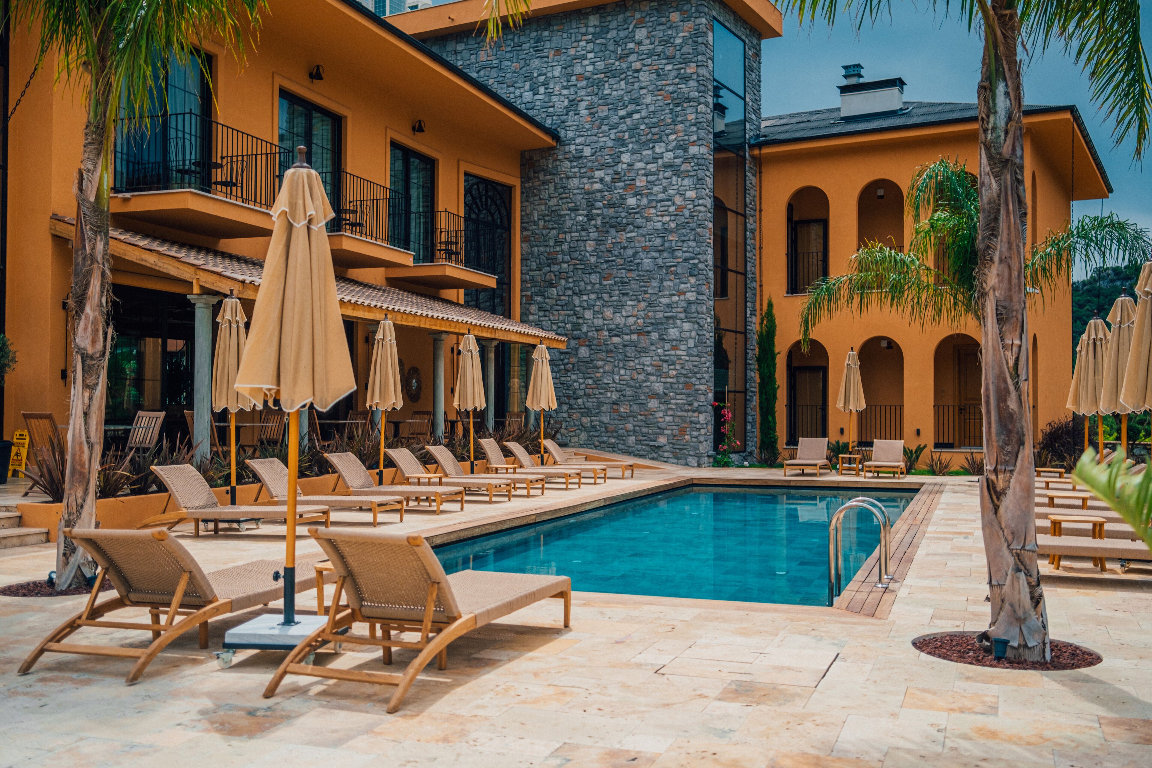 Ein Poolbereich mit Liegestühlen und Sonnenschirmen, ein Steinhaus mit Balkonen und ein klares, blaues Schwimmbecken.