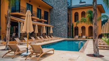 Ein Poolbereich mit Liegestühlen und Sonnenschirmen, ein Steinhaus mit Balkonen und ein klares, blaues Schwimmbecken.